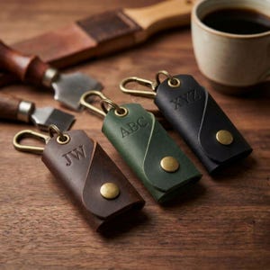 May include: Three leather key organizers in brown, green, and black, each featuring a brass snap button, a metal carabiner clip, and embossed letters. The brown organizer is marked JW, the green one ABC, and the black one is marked XYZ.