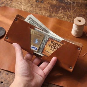 May include: A tan leather wallet featuring four metal rivets at the corners, containing a one hundred dollar bill, a Global Bank Visa card, and a Western Cinema membership card. The wallet is constructed from smooth, light brown leather.
