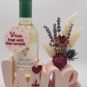 Könnte beinhalten: Eine Flasche Weißwein mit grünem Verschluss steht in einem rosa "Wine Time"-Halter, akzentuiert mit einem kleinen roten Herz. Ein Anhänger mit deutschem Text hängt an der Flasche. Ein kleines Blumengesteck ist ebenfalls vorhanden.