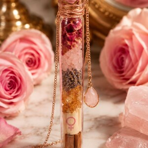 Peut inclure: Un flacon en verre transparent rempli de couches de fleurs séchées, de cristaux et d'herbes, scellé avec un cachet de cire rose. Une chaîne dorée avec un pendentif en quartz rose en forme de goutte pend du flacon. Le fond présente des roses roses et une surface en marbre.