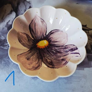 Könnte beinhalten: Eine gewellte Keramikschale mit cremefarbener Basis, die eine handgemalte violette Blume mit leuchtend gelbem und orangefarbenem Zentrum zeigt. Der Innenrand ist mit kleinen, gleichmäßig verteilten goldenen Punkten verziert, die Zahl eins ist blau.