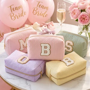 May include: Five rectangular cosmetic pouches in pastel colors including pink, light purple, yellow, and sage green. Each bag features a textured seersucker fabric, a gold zipper, and a large white varsity-style letter patch with a gold glitter border.