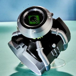 May include: A black and silver wrist-worn device featuring a circular top dial with a black rim. The centre displays a square green screen with a black silhouette of a creature. The base is black with silver accents and curved silver pipes.