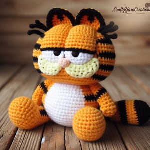 Peut inclure: Une peluche Garfield faite à la main au crochet. Le chat orange a des rayures noires, des accents blancs et un nez rose. La peluche est posée sur une surface en bois. Le texte "Crafty Yarn Creations" est dans le coin supérieur droit.