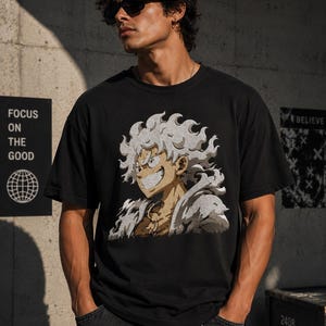 Op de afbeelding: Zwart t-shirt met een cartoonfiguur in wit, beige en geel. Het personage heeft stekelig wit haar en een brede grijns. De persoon draagt een zonnebril.