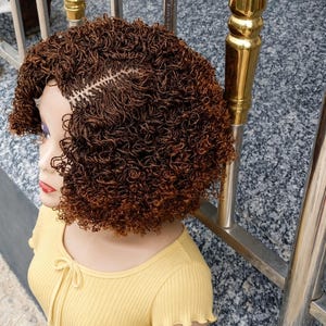 May include: A brown wig with tight, small curls and a visible centre part, styled in a short bob length. The hair features a mix of dark and medium brown tones, creating a textured appearance. The wig is displayed on a mannequin head.