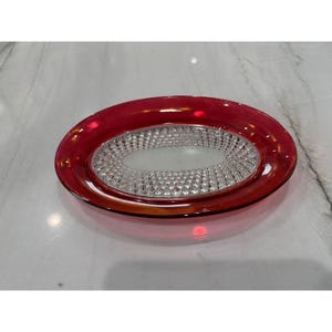 Op de afbeelding: Een ovale glazen serveerschaal met een robijnrode rand en een helder, getextureerd midden. Het midden heeft een diamantachtig patroon. De schaal wordt waarschijnlijk gebruikt om voedsel te serveren of als decoratief stuk.