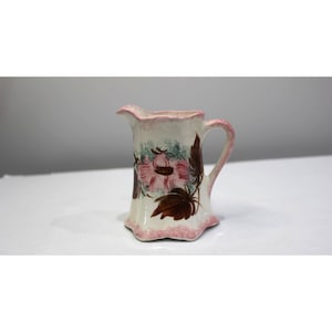 May include: A small, vintage ceramic pitcher with a floral design. The pitcher is primarily white with a pink rim and handle. The floral design features pink flowers and brown leaves. The pitcher has a unique, scalloped base.