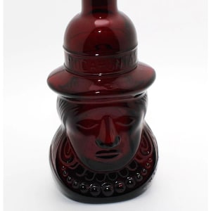 May include: A dark red glass bottle with a detailed face design. The bottle has a hat-like top with the word "CA-ON" embossed on it. The face has a necklace of small, round beads.