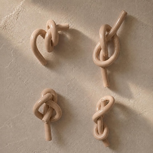 May include: Four individual beige ceramic objects, each shaped like a knotted rope or cord. The pieces feature a smooth, glossy finish and are arranged in a grid pattern. Each object displays a unique, complex knot configuration with rounded ends.