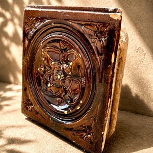 May include: A square, brown ceramic tile featuring a raised floral design. The tile has a glossy finish and is framed by a decorative border with floral accents. The tile is propped against a neutral-toned surface.