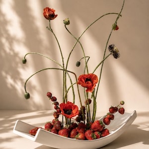 May include: A white, curved, boat-shaped ceramic bowl filled with fresh red strawberries. Several tall, slender green stems rise from the fruit, topped with vibrant orange-red poppy flowers and closed green poppy buds, creating a floral arrangement.