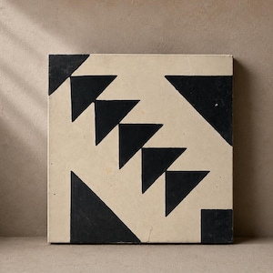 May include: Square tile with a cream-colored background and a black geometric design. The design features triangles and other shapes arranged in a repeating pattern. The tile is likely made of ceramic or a similar material.