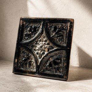 May include: A square, black decorative tile with an ornate, raised floral design. The tile features a central floral motif surrounded by four quadrants, each with a stylized leaf pattern. The tile has a distressed, aged appearance.