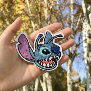 May include: An embroidered patch featuring the head of a blue alien character with large ears, a wide grin showing white teeth, and a pink tongue. The character has light blue skin, dark blue accents, and a pink inner ear, all outlined in white thread.