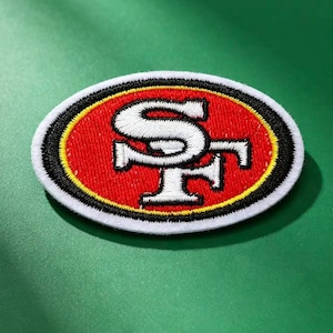 Puede incluir: Un parche bordado de forma ovalada con fondo rojo y un borde blanco y negro. El centro muestra las letras blancas SF en una fuente estilizada, delineadas en negro, que representan el logotipo del equipo deportivo San Francisco 49ers.