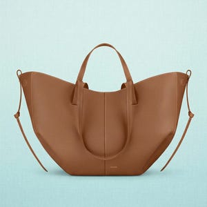 May include: A brown leather tote bag with two short handles and two long shoulder straps. The bag has a wide opening and a structured design. The stitching is visible, and the bag has a minimalist aesthetic.