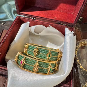 Op de afbeelding: Twee goudkleurige armbanden met rijen kleine groene kralen. Elke armband is versierd met gouden metalen motieven, waaronder kleine figuren en marquisevormige inzetstukken met elk een enkele groene of roze steen.
