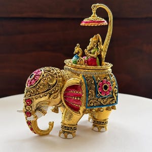 May include: A gold-toned decorative elephant figurine featuring intricate filigree patterns, pink and blue enamel accents, and white tusks. A small deity figure sits on the elephant's back under a golden canopy with a pink underside.