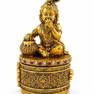 May include: A gold-toned metal figurine of a child sitting on a cylindrical box. The child has a peacock feather with a green stone on their head and holds a small pot. The box features intricate floral engravings and a border of red and white stones.
