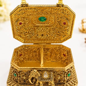 May include: An ornate gold-toned metal jewelry box with an open hinged lid. The exterior features intricate filigree patterns, an embossed elephant figure, and inlaid green, red, and clear stones. The interior is divided into two sections.