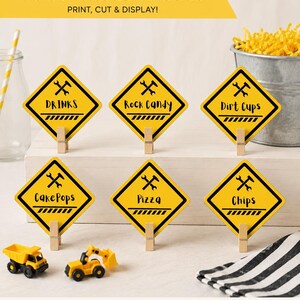 May include: Yellow food table labels with black text and construction-themed graphics. Labels include "Drinks," "Rock Candy," "Dirt Cups," "Cake Pops," "Pizza," and "Chips." Each label is held by a wooden clip. The text "FOOD TABLE LABELS" is at the top.