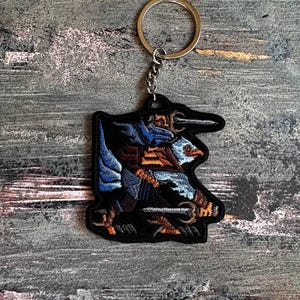 Op de afbeelding: Zwarte geborduurde sleutelhanger met een gedetailleerd ontwerp met blauwe, bruine en witte accenten. De sleutelhanger heeft een zilveren metalen ring en ketting. Het ontwerp toont een gestileerde vogel of mythisch wezen.