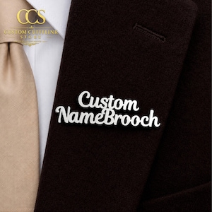May include: A silver-colored "Custom Name Brooch" pin is displayed on a dark brown blazer. The pin is in a cursive font. A tan tie and white shirt are visible in the background. The top left corner shows the logo of "Custom Cufflink Store".