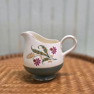 May include: A vintage cream pitcher with a white upper section and a teal base. The pitcher features a floral design with purple flowers, green leaves, and gold accents. It has a curved spout and a rounded handle.