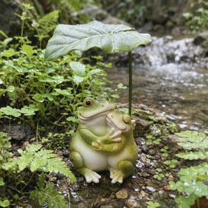 May include: A charming green frog ornament embracing a smaller frog, sheltered under a large leaf. The frogs are positioned near a stream, surrounded by lush greenery. The ornament is a delightful decorative item.