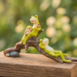 May include: Two whimsical frog figurines in shades of yellow and green are perched on a piece of driftwood. One frog is playing a musical instrument, while the other relaxes. The figurines rest on a wooden surface against a blurred green background.