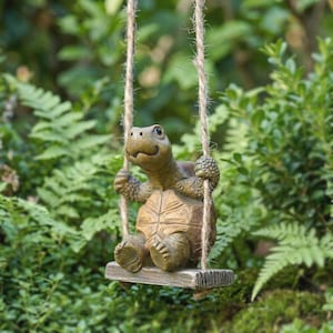 Puede incluir: Un adorno de jardín caprichoso que presenta una tortuga sonriente en un columpio. La tortuga, con un caparazón detallado y una expresión amigable, está suspendida de un columpio de madera por una cuerda. La estatua está situada sobre un fondo de exuberante follaje verde.