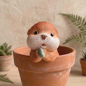 May include: A ceramic otter figurine with a surprised expression, holding a green leaf, peeks out from a terracotta flower pot. The otter has brown fur and white cheeks. The pot is empty, and other small potted plants are in the background.