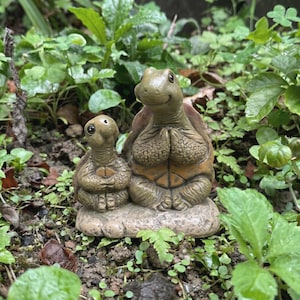 May include: Two ceramic turtle ornaments, a larger one and a smaller one, positioned together on a stone base. The turtles are green and brown with detailed shell patterns. The background shows vibrant green foliage.
