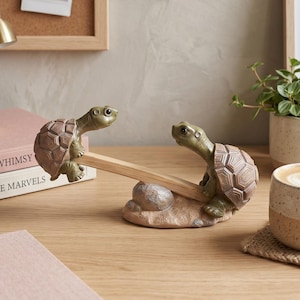 May include: A decorative desk tidy featuring two turtle figurines in green and brown, balancing a wooden pencil. The turtles are positioned on a small, textured base, adding a playful touch to a desk or workspace. The pencil is made of wood.