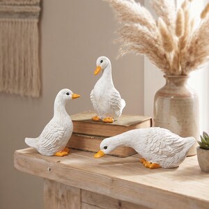 May include: A set of three white duck figurines with orange beaks and feet. One duck stands upright on a stack of two brown books, another stands to the left, and the third is positioned in a forward-leaning posture on the wooden surface.