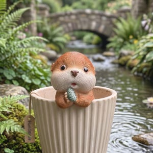 May include: A ceramic otter figurine with brown fur and a surprised expression, holding a small green leaf. The otter is inside a beige, ribbed planter. A small tag reads "Handmade". The background features a stone bridge and a flowing stream.