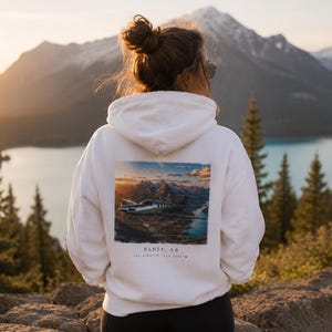 Peut inclure: Un sweat à capuche blanc avec une image d'un avion survolant un lac et des montagnes au coucher du soleil. Le texte "BANFF, AB" et les coordonnées sont imprimés en dessous. La personne se tient sur une surface rocheuse.