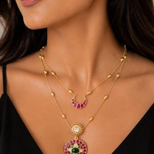 Pode incluir: Um conjunto de colar em camadas de tom dourado com um pingente em forma de meia-lua com pedras vermelhas e um pingente circular com uma pedra verde central rodeada por pedras vermelhas e brancas, com brincos quadrados a combinar.