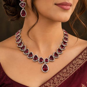 Puede incluir: Un conjunto de joyería compuesto por un collar y pendientes a juego. El collar presenta una serie de piedras de color rojo intenso en forma de pera rodeadas de pequeños cristales blancos, con un colgante central más grande.
