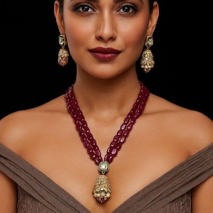 May include: A two-strand necklace featuring smooth, deep burgundy oval beads and a central gold-toned pendant with intricate filigree and small white pearls. Matching bell-shaped drop earrings with gold-toned filigree and white pearl accents.