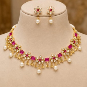 May include: A gold-toned necklace and matching earring set featuring flower-shaped motifs with central fuchsia rectangular stones, clear accent crystals, and dangling white pearls. The necklace has a repeating floral pattern with gold beads.