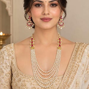May include: A multi-strand pearl necklace featuring five layers of small white pearls with gold-toned connectors and teardrop-shaped pink stones. Matching chandelier-style earrings feature a circular gold frame with pink stones and dangling pearls.