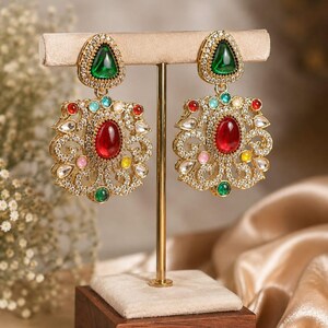 May include: A pair of gold-tone dangle earrings featuring a triangular green stone top and a large ornate pendant base. The pendant is encrusted with clear crystals, a central oval red stone, and smaller accent stones in red, blue, pink, and yellow.