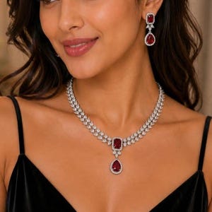 Pode incluir: Um conjunto de colar e brincos prateados com pedras vermelho-escuras retangulares e em forma de pera. O colar consiste em duas filas de pequenas pedras transparentes com uma pedra vermelha retangular central e uma pedra vermelha pendente.