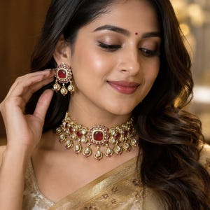 May include: A gold-toned choker necklace and matching stud earrings featuring deep red square-cut stones surrounded by small white crystals. The jewellery set includes dangling teardrop-shaped accents with red and white stones in a metal setting.