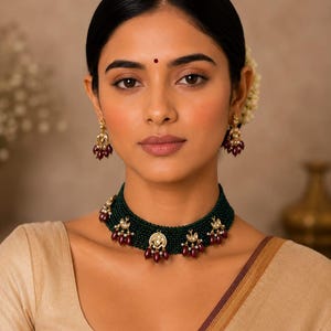 Può includere: Una collana girocollo composta da più fili di piccole perline verde scuro, arricchita da ciondoli in metallo dorato e perline bordeaux a forma di goccia, abbinata a orecchini pendenti coordinati che riprendono gli stessi dettagli oro e bordeaux.