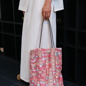 May include: A rectangular tote bag featuring a textured surface covered in overlapping pink, peach, and iridescent silver sequins arranged in a scalloped pattern. The bag includes two thin, dark grey shoulder straps attached to the top edge.