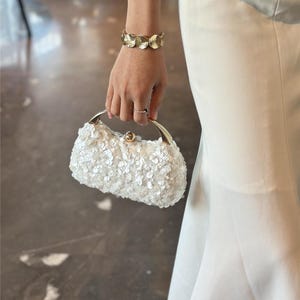 May include: A white, sequined clutch with gold-tone hardware is held by a person. The clutch has a rounded shape and a small, gold clasp. A gold bracelet with circular accents is worn on the wrist. The background is blurred, with a glimpse of a white dress.