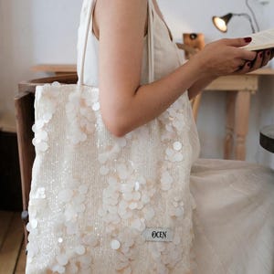 Peut inclure: Un sac fourre-tout blanc doté d'une surface texturée recouverte de nombreuses paillettes blanches rondes et semi-translucides. Le sac comprend deux bandoulières et une petite étiquette blanche rectangulaire avec le texte OCEN en bas à droite.